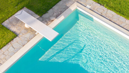 Aerial view of a rectangular swimming pool with diving board, belonging to a large villa. The water is transparent and through the blue you can see the steps. Around the water there is a stone floor.