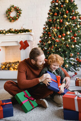 Obraz na płótnie Canvas cheerful redhead boy holding Christmas present while sitting on floor near bearded father in decorated living room
