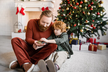 Obraz na płótnie Canvas redhead father and child holding Christmas bauble on floor in living room with festive decor