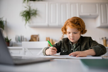 redhead child holding pen while learning at home near blurred laptop