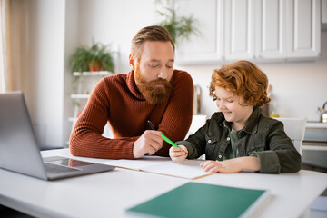 Fototapeta premium bearded man assisting smiling redhead son writing in notebook near blurred laptop
