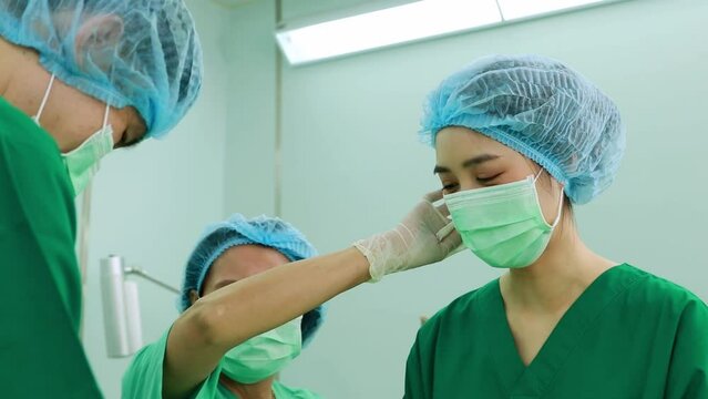 Senior female nursing assistant using sweat towel to wipe the female surgeon's face during the operation of the patient of the professional medical team in the operating room with determination.