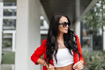 Beautiful fashion business woman with fashionable sunglasses in stylish casual clothes with red blazer, white top and purse walking in the city near a modern building