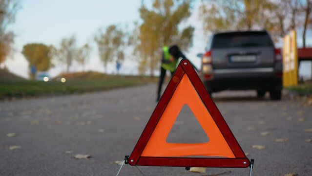 Reflecting warning triangle on road behind broken car and driver inspect rear wheel. Emergency stop sing on side of road
