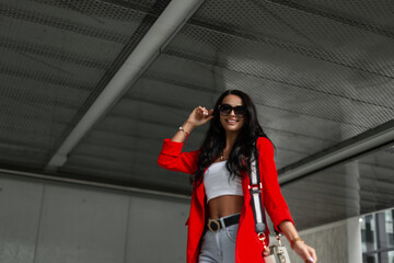Happy stylish beautiful fashionable business girl with smile wears cool trendy sunglasses in a fashion red blazer, top and jeans with bag walking in the city