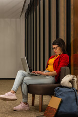 Young casually dressed traveler woman in waiting room sitting on a chair typing on her laptop while supporting it on her legs