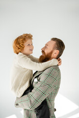 Trendy father in jacket hugging and lifting cheerful redhead son on grey background