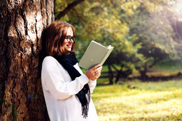 Young adult woman in glasses and scarf reads book leaning on the trunk of tree in the park and smiling.