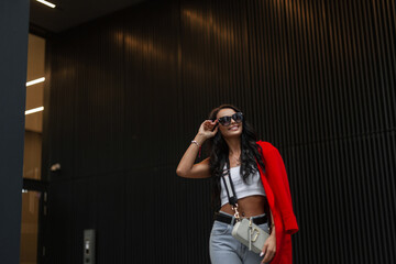 Fashion beautiful business girl in stylish casual clothes with top, jeans, red jacket and bag walking in the city and wearing stylish sunglasses near a modern building