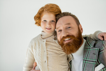 Portrait of positive man in jacket hugging son and looking at camera isolated on grey