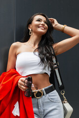 Pretty young beautiful smiling happiness woman with smile in fashion business clothes with top, jeans, jacket and handbag stands near a merl black wall