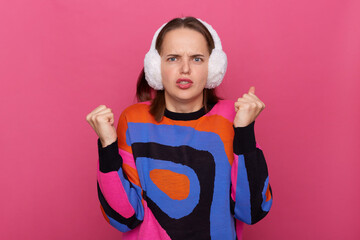 Photo of angry aggressive Caucasian woman wearing fluffy fur earmuffs and warm sweater standing isolated over pink background with clenched fists, looks stressed, arguing with someone.