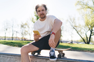 Young man with smartphone sitting on skateboard in skate park