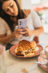 Chica joven guapa morena desayunando croissant relleno en cafeteria estilo cozy