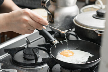 detailed view of a girl's hand holding a spoon to pour the oil over the egg that is frying in a small frying pan.