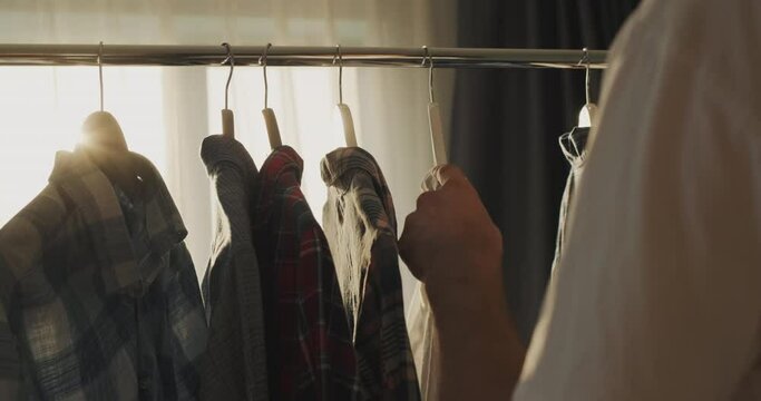 A man chooses his clothes near a rack with a coat hanger, from behind he is illuminated by the sun's rays from the window. Rear view