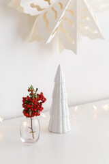 Christmas cozy winter home decor. New year interior decoration. Branch with red berries in vase, decorative ceramic house and christmas tree, glowing garland lights. Stylish composition on the dresser