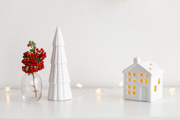 Christmas cozy winter home decor. New year interior decoration. Branch with red berries in vase, decorative ceramic house and christmas tree, glowing garland lights. Stylish composition on the dresser