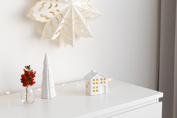 Christmas cozy winter home decor. New year interior decorations. Paper snowflake on wall, branch with red berries in vase, decorative ceramic house and christmas tree. Stylish composition on dresser.