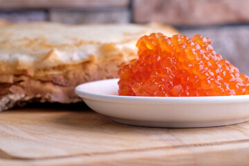 red caviar in a white porcelain bowl next to a stack of thin flour pancakes stands on a wooden stand