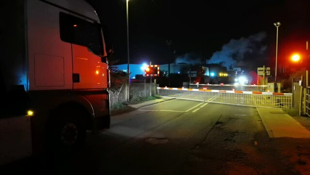 A lorry waits at a railway level crossing by safety barriers as a freight train goes past at night. Warning lights flash as a train approaches.