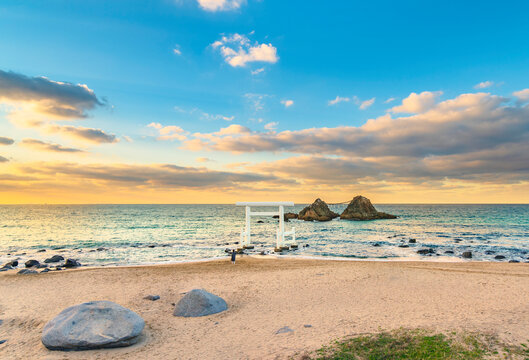 One person praying on the Itoshima Beach of Fukuoka standing in front of a white Shinto torii gate leading to the famous Sakurai Futamigaura's Meoto Iwa Couple Rocks in the evening sunset light.