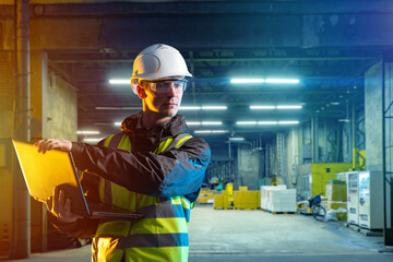 Logistics specialist. Man with laptop. Guy near entrance to warehouse. Worker in yellow vest and protective helmet. Man employee logistics company. Concept providing logistics services. Art blurred