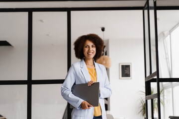 Cheerful african young woman programmer is standing with laptop and smiling. Candid african american girl with laptop standing in office, smiles and rejoices at successful work in IT company.