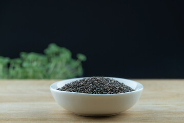 Still life black seeds chia in white plate on wooden platform. Micro greens plant salvia hispanica grow on black background. Germination of herb cereal seeds. Healthy nutrition mineral organic food.