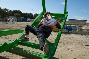 Fototapeta premium a handsome stylish fair-haired boy posing among green colored iron structures in a knitted sweater and jeans against a bright blue cloudy sky