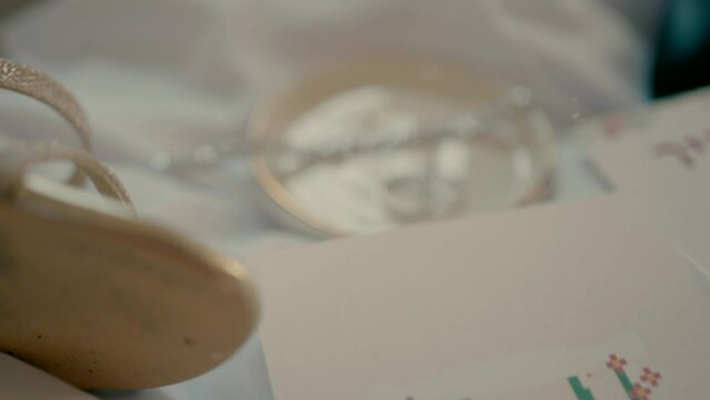 The bride's bracelet and rings in a ceramic saucer with bridal shoes next to it on satin. A close-up shot of a bride's things and an invitation on a wedding day.
