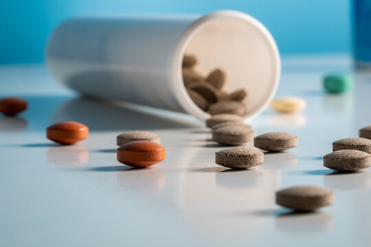 Round brown pills spilled out of bottle onto a glossy, reflective surface. Close up of dosed medical drugs, vitamins, antibiotics on blue background. Medicinal substances for treatment of coronavirus