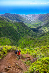 Fototapeta premium hiker girl climbs steep trail on oahu with honolulu skyline in background on wiliwilinui ridge trail; hiking on green mountains in hawaii, holiday in hawaii