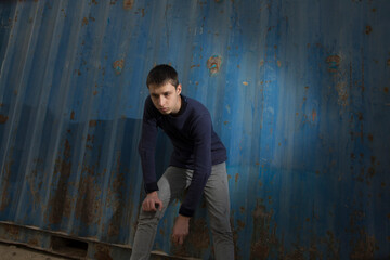 Obraz na płótnie Canvas young caucasian guy posing with a serious face dressed in a sweater and jeans in an industrial area against a blue iron wall and a blue cloudy sky. lower angle