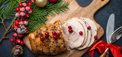 Christmas baked ham sliced with red berries and festive decorations on wooden cutting board, dark rustic background from above. Christmas and New Year holiday dinner with baked pork 