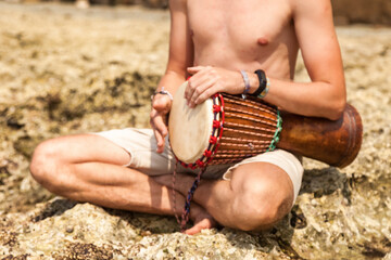 Close up young man drummer, playing rhythm with djembe drum, sitting at tropical coral reef coast...