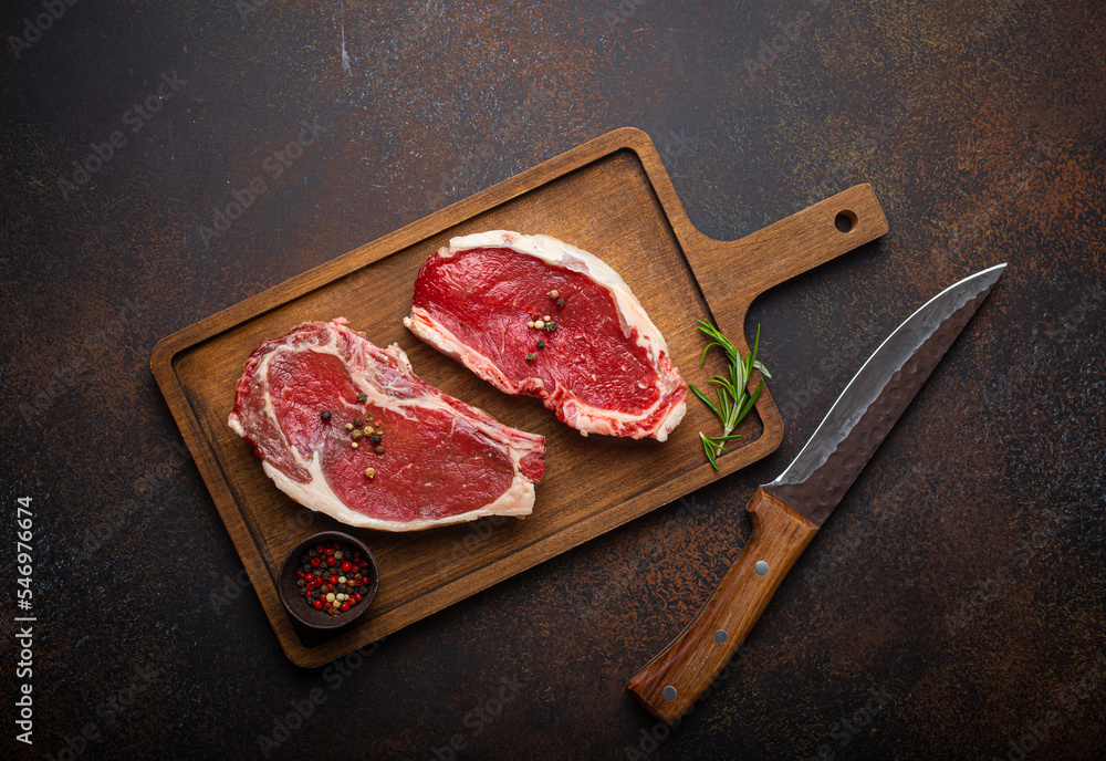 Wall mural Two raw uncooked meat beef rib eye marbled steaks on wooden cutting board with seasonings on dark rustic background ready to be grilled from above, preparing dinner with meat