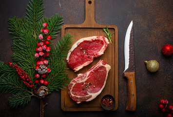 Two raw uncooked meat beef steaks on wooden cutting board with knife and seasonings on dark rustic background with Christmas festive decoration from above 
