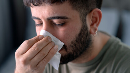 Unhappy sad young indian male in  suffering from fever and flu on sofa, blowing nose in napkin in bedroom interior. 