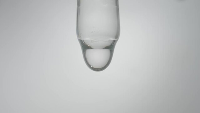 Drop of transparent liquid, oil, serum or tincture dripping from glass pipette on a gray background. Macro shot of dripping drops of clean essential oil or medicinal solution. Concept of medicine.