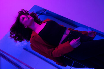 Sensual brunette young model in red suit and black jeans in mixed pink and violet neon light, posing down on mirror on the floor.