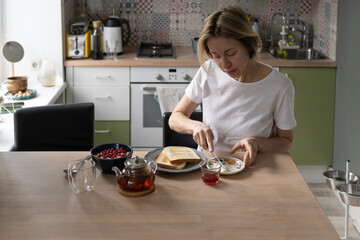 Mature woman sitting at table in kitchen makes breakfast. Daylight breaks through window...