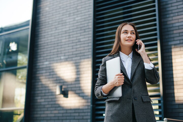 Portrait of an attractive business woman with laptop walking on city street and making a call on...