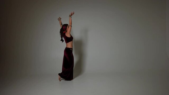 Side view slim woman with wing tattoo on arm raising hands performing at grey background in slow motion. Wide shot of Caucasian talented belly dancer dancing Middle Eastern dance