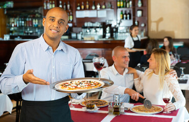 Fototapeta na wymiar Smiling waiter holding serving tray with pizza at restaurant with customers his behind