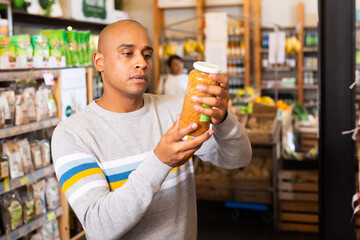 Interested Latin American man reading product label on jar while choosing groceries in supermarket