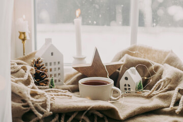 Warm cup of tea, candles, lights, little christmas house and tree, wooden star on cozy blanket on windowsill. Winter hygge. Atmospheric Christmas background. Cozy home on snowy day