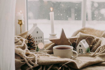 Winter hygge. Warm cup of tea, candles, lights, little christmas houses and trees, wooden star on cozy blanket on windowsill. Cozy home. Atmospheric snowy day. Christmas background