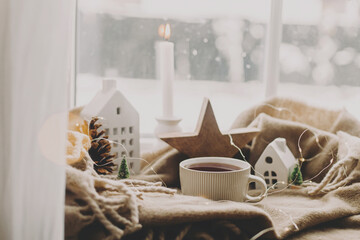 Warm cup of tea, candles, lights, little christmas house and tree, wooden star on cozy blanket on windowsill. Winter hygge. Atmospheric Christmas background. Cozy home on snowy day - obrazy, fototapety, plakaty