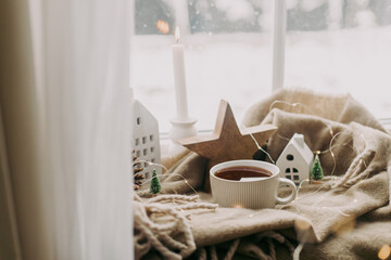 Cozy home at snowy day. Warm cup of tea, candles, lights, little christmas houses and trees, wooden...